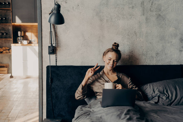 Piękny radosny młoda dziewczyna leżąca w łóżku po przebudzeniu w godzinach porannych, mając filiżankę gorącej kawy podczas rozmowy wideo z przyjaciółmi online na laptopie, uśmiechając się i pokazując znak V palcami - Zdjęcie, obraz