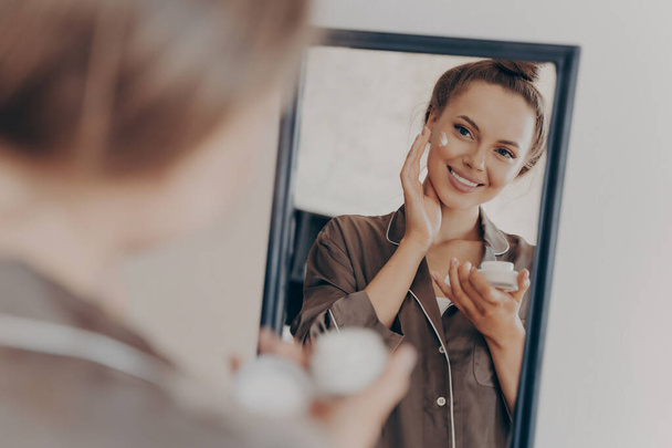 Refleksja w lustrze pięknej kobiety w jedwabnej piżamie uśmiechniętej podczas stosowania kremu do twarzy dla głębszej naprawy, ochrony skóry twarzy. Pielęgnacja skóry, piękno i skuteczne leczenie zmarszczek koncepcja - Zdjęcie, obraz
