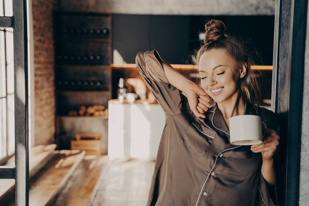 New day. Happy beautiful brunette female stretching with cup of coffee in her hand enjoying morning sunshine while standing in kitchen doorway in satin pajama. People and lifestyle concept - Foto, Bild