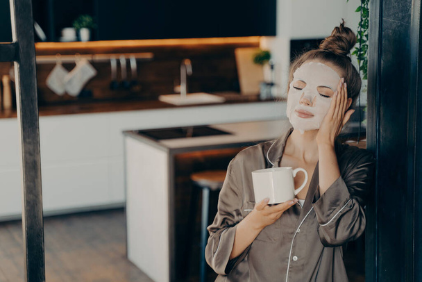 Mujer joven en pijama de seda que se queda en casa y disfruta de la taza de café de la mañana mientras se aplica la máscara desechable de cuidado de la piel blanca en la cara. Procedimiento de rutina diaria para el rejuvenecimiento y cuidado de la piel - Foto, imagen