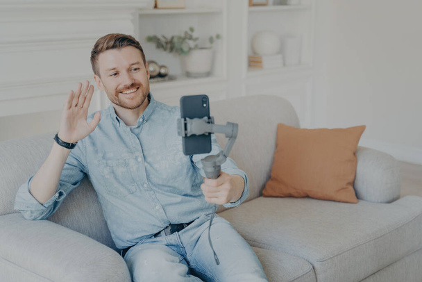 Молодой человек наслаждается видеочатом с друзьями, смотрит на экран телефона и машет рукой, веселый бородатый мужчина держит смартфон со стабилизатором подмышки в руке, сидя на диване в гостиной - Фото, изображение