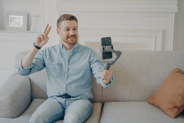 Young man with short beard wearing casual clothes and sitting on comfortable couch, video chatting holding gimbal with phone attached to it, showing peace gesture to camera - Fotografie, Obrázek