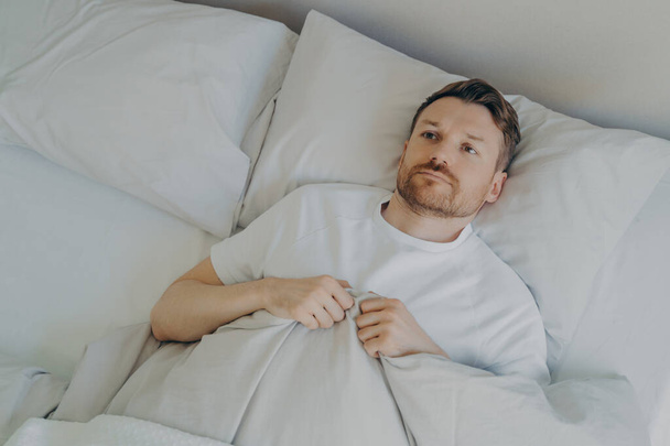 View from above of lonely upset young bearded man lying in bed with open eyes and can't sleep, feeling unhappy and tired, squeezes blanket with hands and being stressed of insomnia. Sleeping problems - Fotoğraf, Görsel