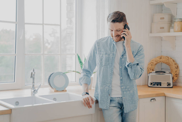 Handsome busy bearded businessman in casual comfy clothes standing in kitchen holding mobile phone, talking with client and dealing with customer issues while working remotely from home - Photo, Image