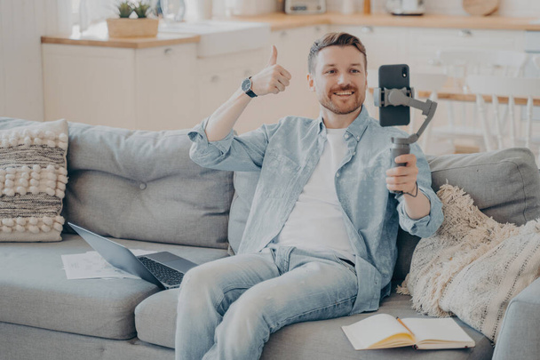Confident young male freelancer showing thumbs up gesture to his client to inform him that everything is going well through phone video call, sitting on couch holding gimbal with phone - Foto, afbeelding