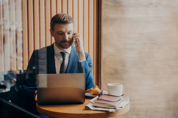 Portrait of elegant financier in stylish suit sitting in cafe with open laptop in front of him while talking on cellphone about work and business plan with concentrated look, verifying info on screen - Photo, Image