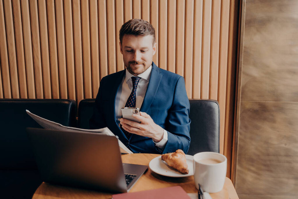 Confident young businessman in formal clothes enjoying coffee and trying to keep up with busy work schedule on his smartphone, working on laptop and reading newspaper while sitting at coffee shop - Photo, Image