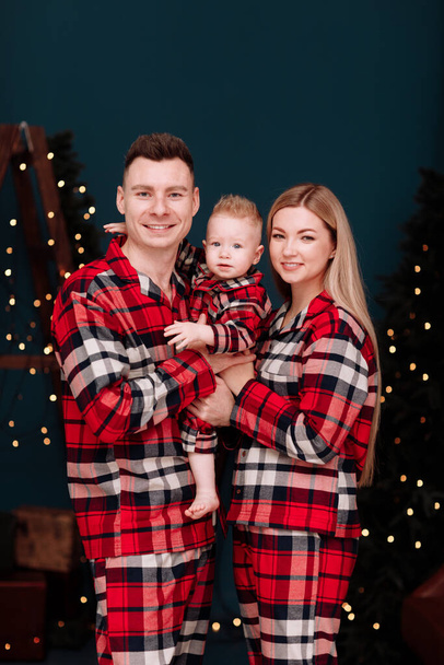 Christmas Family holidays. Smiling young dad, mom are hugging baby boy son in the same red checked pyjamas near the decorations Christmas tree with light at home. Happiness. New Years concept. - Zdjęcie, obraz