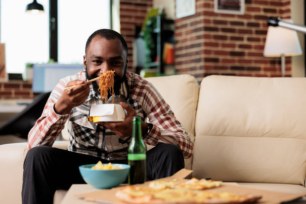 African american man using chopsticks to eat takeout noodles food at home, watching movie on tv channel. Eating takeaway delivery meal from box and enjoying film on television. - Photo, image