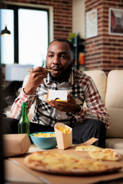 Relaxed man enjoying noodles package from takeaway delivery, eating food with chopsticks in front of television movie. Watching film on tv channel program, having fun with takeout meal. - Photo, image