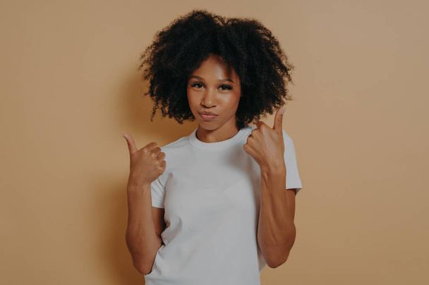 Success sign. Confident young african woman showing thumbs up with both hands and looking at camera isolated on beige background, showing winner gesture and smiling. Body language concept - Photo, Image