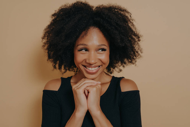 Studio shot of happy Afro girl keeps hands together under chin, being in high spirit, likes friends idea for next day, isolated on dark beige wall background with copy space. Positive emotions concept - Photo, image