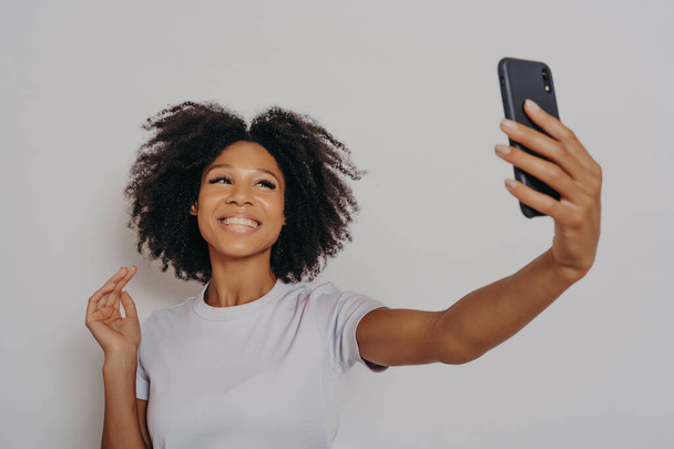 Pozitivní veselé tmavé pleti žena fotografování na moderní smartphone kamery, veselá africká žena oblečená v ležérní bílé tričko přičemž selfie na mobilním telefonu, šťastně se usmívá - Fotografie, Obrázek
