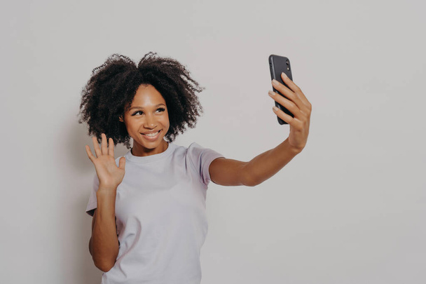 Gelukkige jonge vrouw zwaaien op de camera tijdens het hebben van video call met de beste vriend op de moderne smartphone, jonge vrolijke Afrikaanse vrouw uiten positiviteit tijdens het praten met volgers in sociale media - Foto, afbeelding