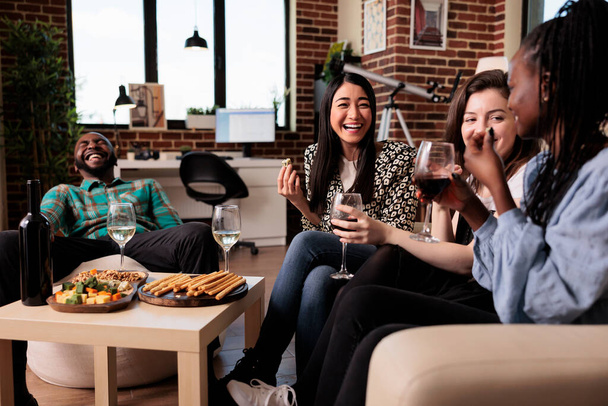 Close friends having wine party and laughing with mouth wide open while celebrating friendship event. Diverse group of people in living room enjoying relaxing time together while bonding. - Foto, afbeelding