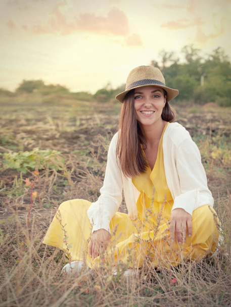 Beautiful young woman portrait sitting on sunset evening field, smiling woman dressed in yellow dress have a rest outdoor getaway - Фото, изображение