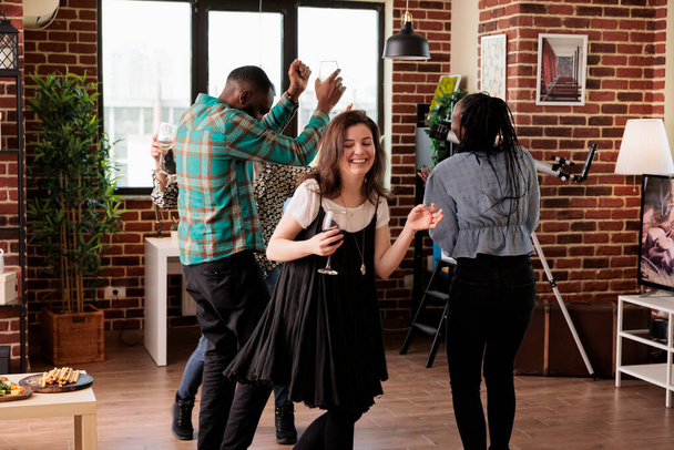 Multiethnic group of closed friends dancing together in living room at wine party while having fun. Young adult people enjoying disco music at home while celebrating friendship anniversary event. - Foto, Imagen