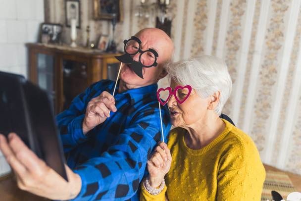 Cute joyful senior European couple holding party accessories or masks on wooden sticks - fake black mustache and pink heart-shaped glasses, laughing and taking selfies on a phone, enjoying retirement - Foto, afbeelding