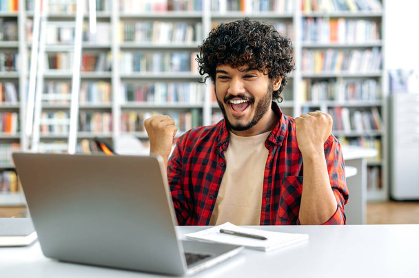 Amazed happy excited stylish mixed race guy, student, sitting in university library with laptop, rejoices in success, win, good mark on the exam, gesturing hands, smiling, emotional face expression - Photo, image