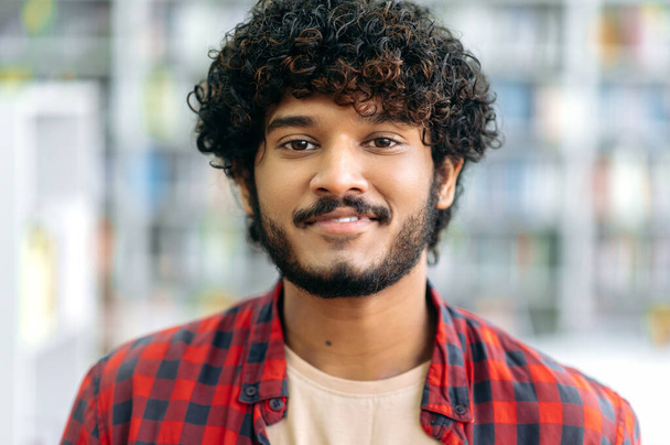 Close-up photo of a handsome positive curly haired indian or arabian guy, in casual shirt, manager, designer, student or freelancer, stands indoors, looks at camera, smiling friendly - Foto, Imagen