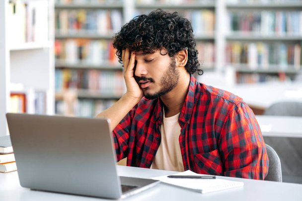 Tired student of Arabian or Indian nationality falling asleep at a desk in the library, in stylish clothes, tired of preparing for the exam, reading books, feels exhausted, closed his eyes, needs rest - Foto, imagen