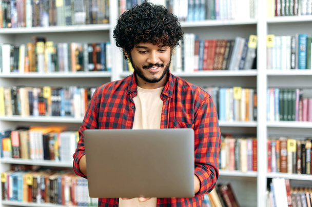 Stylish modern curly haired positive arabian or indian university student, holding an open laptop in his hands, standing in the library, browsing the internet, looking for information, smiling - 写真・画像
