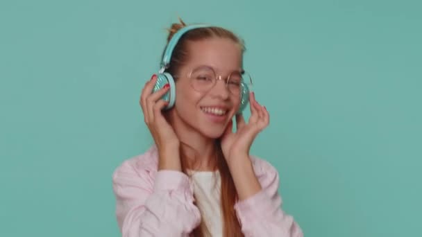 Happy pretty teenager relaxed girl in glasses listening music via headphones and dancing disco fooling around having fun expressive gesticulating hands. Young stylish teen female child kid indoors - Filmagem, Vídeo