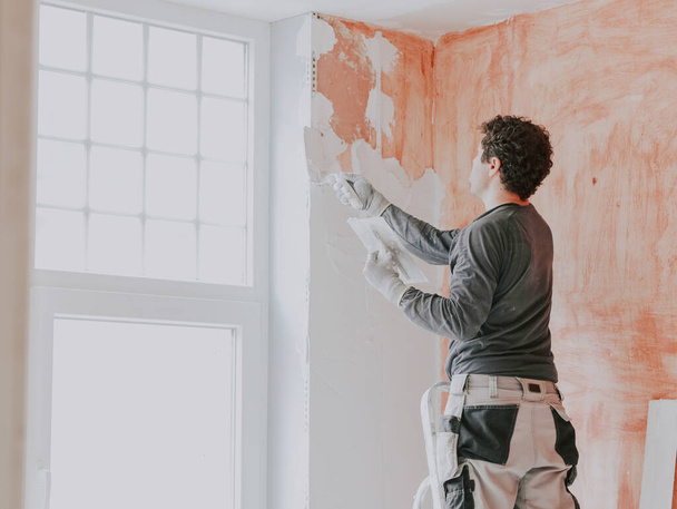 A young caucasian guy in a gray t-shirt with sleeves, work pants and curly brown hair applies fresh putty with a small spatula on the wall near the window, standing on a stepladder, close-up side view. The concept of apartment and house renovation, w - Photo, image