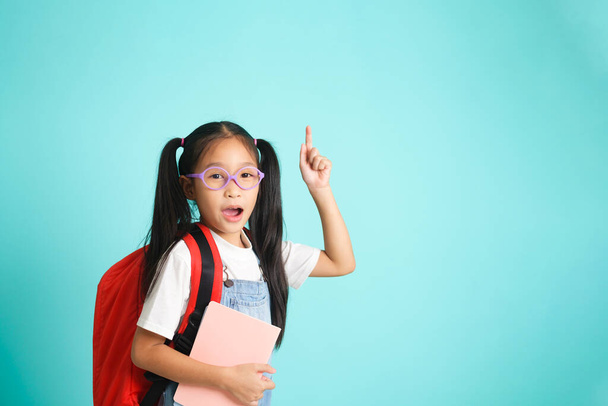 Χαμογελαστό κοριτσάκι με γυαλιά που δείχνει το δάχτυλο προς τα πάνω πηγαίνοντας σχολείο, σχολική ιδέα. στέκεται σε μπλε φόντο. - Φωτογραφία, εικόνα
