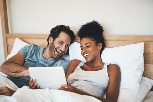 Happy, relaxed and carefree couple reading social media news on digital tablet and laughing in bed. Interracial husband and wife waking up together and browsing internet, sharing a funny online joke. - Photo, image