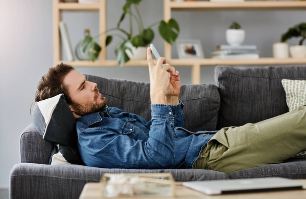 Ξάπλωσε, χαλάρωσε και μείνε επίκαιρος. ένας όμορφος νεαρός χρησιμοποιώντας ένα smartphone, ενώ βρίσκεται σε έναν καναπέ στο σπίτι - Φωτογραφία, εικόνα