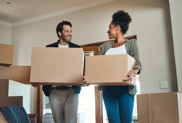 Счастливая пара или новые домовладельцы переезжают вместе, перевозя коробки с мебелью или вещами и имуществом. Любящие, межрасовые партнеры после покупки недвижимости, улыбающиеся друг другу - Фото, изображение