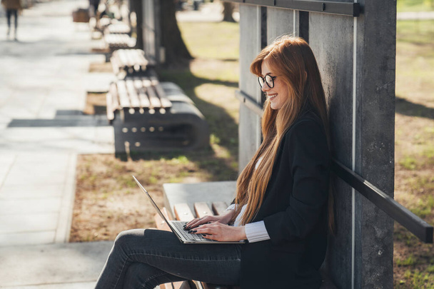 Side view of a ginger business woman freelancer sitting on bench working with laptop in city park. A beautiful happy woman using laptop outdoors. Distance - Zdjęcie, obraz