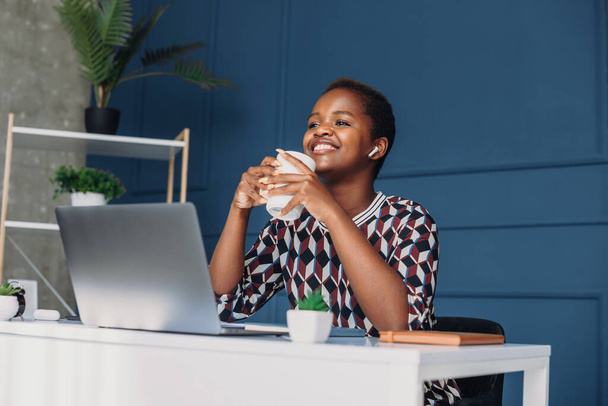 Χαμογελώντας μια Αφροαμερικανή που απολαμβάνει τον πρωινό καφέ, κάθεται στο γραφείο στο γραφείο. Απομακρυσμένη δουλειά. Τεχνολογία Διαδικτύου. - Φωτογραφία, εικόνα