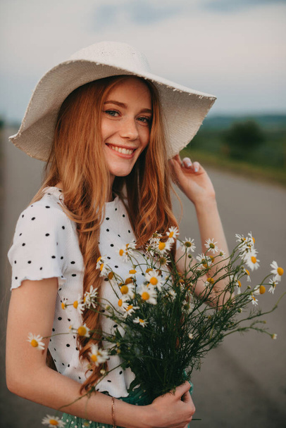 Портрет симпатичной привлекательной рыжеволосой кавказской веснушки в соломенной шляпе с букетом цветов. Деревенская жизнь. Солнечный день. Летняя природа. - Фото, изображение