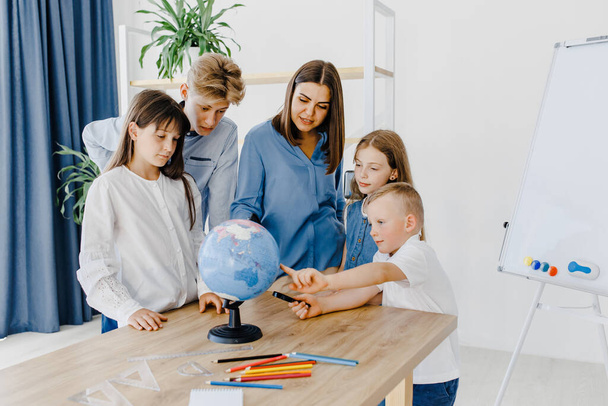 Teacher and children in class are looking at globe, teacher helps explain the lesson to the children in the class at a desk. Educational school process, bright room and interesting learning - Zdjęcie, obraz