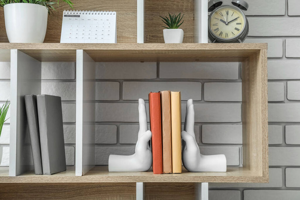 Bookcase with holder, calendar, alarm clock and flowerpot near white brick wall - 写真・画像