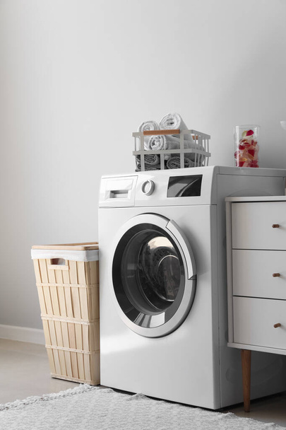 Стиральная машина с корзиной, полотенцами и капсулами возле светлой стены в прачечной - Фото, изображение