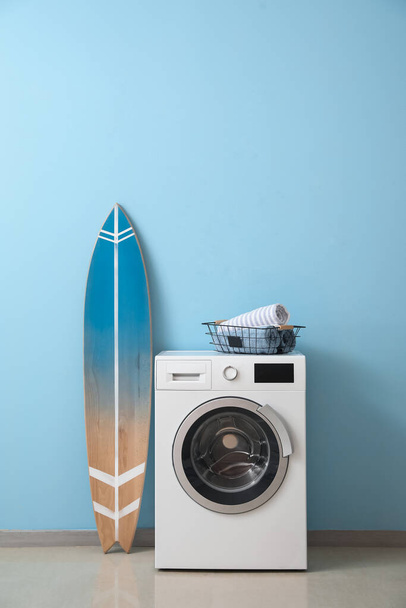 Корзина с полотенцами на стиральной машине и доска для серфинга возле синей стены - Фото, изображение