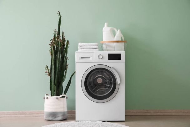 Πλυντήριο ρούχων με καλάθι, απορρυπαντικό, πετσέτες και κάκτους κοντά σε πράσινο τοίχο - Φωτογραφία, εικόνα