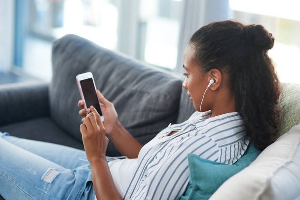 Searching for some new shows to stream on her cellphone. a young woman wearing earphones while using a cellphone at home - Foto, imagen