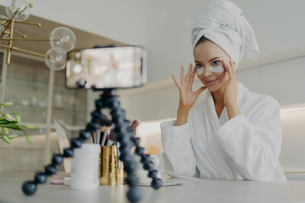 Young beautiful woman blogger in bathrobe recording video tutorial for beauty blog or social media how to use cosmetic patches or pads from dark circles while sitting in kitchen. Skin care concept - Photo, Image
