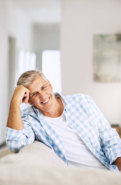 Casual, relaxed and happy mature man smiling indoors on his couch at home on a relaxing day. Portrait of a handsome senior male sitting on his comfortable sofa enjoying his time alone at his house. - Photo, Image