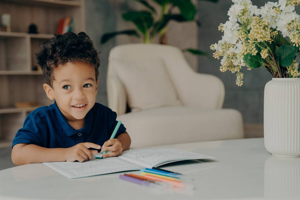 Boldog göndör kevert verseny fiú színező állatok kifestőkönyv filctollal, félrenéz és mosolyog, miközben ül az asztal előtt kényelmes nappaliban otthon. Szabadidős tevékenységek gyermekeknek - Fotó, kép