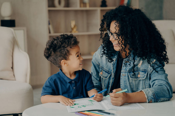 Beautiful afro american family of two, mother and son looking at each other while spending time at home by painting book with different colors in living room. Motherhood and parenting concept - Photo, Image