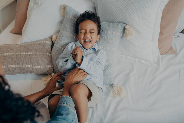Woman mom tickles adorable mixed race little son with curly hair lying on pillows in bed. Cute baby boy in light colored casual clothes having fun and sincerely laughing. Family weekend leisure time - Fotó, kép