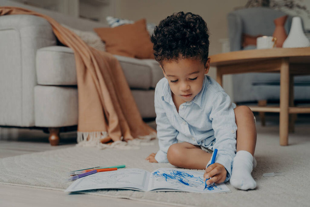 リビングルームの快適なカーペット敷きの床に座っている小さな焦点を当てたアフリカの赤ちゃんの少年は、着色本に青い鉛筆で描画します。家庭での子供の余暇と子供のための創造的な趣味の概念 - 写真・画像
