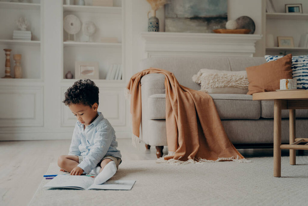 Mulatto curly haired cute boy calmly sitting on warm floor playing with pencils in stylishly furnished living room with sofa and coffee table beside him, creative child activity indoor concept - Foto, imagen