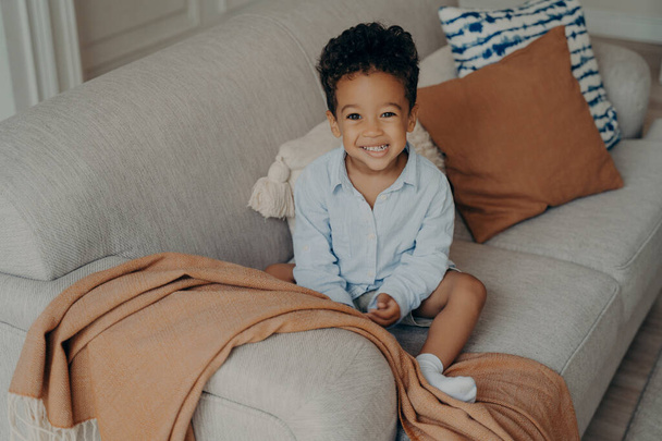 Υπέροχο αφρο-αμερικάνικο παιδί με σγουρά μαλλιά και χαριτωμένο χαμόγελο κάθεται στον καναπέ με μπεζ χρωματιστά στοιχεία μαξιλάρια και κάλυμμα, κοιτάζοντας ψηλά στην κάμερα και χαμογελώντας ενώ παίζει στο σαλόνι στο σπίτι - Φωτογραφία, εικόνα