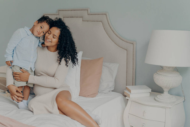 My little boy. Happy pregnant afro american woman in casual white dress with curly hair sitting on bed with her sweet little son and showing strong feelings of love and care. Maternity and pregnancy - Photo, Image